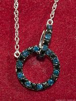Close-up front view of the silver circular pendant with blue and black stones set into it, attached to a silver chain, displayed on a red velvety background.
