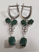 Front view of silver drop earrings with three oval emeralds and clusters of cubic zirconia stones