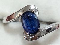 Close-up front view of silver ring with oval blue sapphire set in a prong setting, showing the swirl band design and sapphire color.