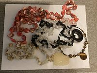 Photo of four different beaded necklaces of various colors and materials on a white surface, showing details of beads and pendants.