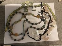 Photo showing multiple beaded necklaces and pendants made from semi-precious stones and shell discs on black cord.