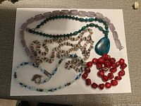 Photo showing a collection of necklaces and a bracelet with various semi-precious stones and glass beads in different shapes and colors arranged on a white sheet.