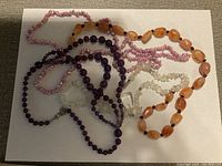 Four semi-precious stone beaded necklaces in various colors and bead shapes laid out on a white surface.