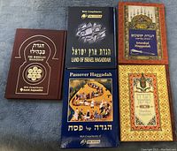 Photo of five Jewish Haggadah books arranged on a surface showing varied covers, colors and titles.