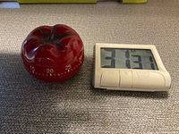Photo shows two kitchen timers side by side on a beige surface. The timer on the left is a red tomato-shaped mechanical timer. The timer on the right is a white digital timer with LCD display showing time and three buttons below the screen.