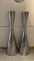 Two tall, stainless steel candle holders with a modern hourglass shape shown standing side by side against a cardboard background.