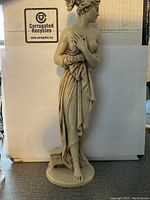 Front view of the beige sculpture of Goddess Aphrodite Venus showing draped fabric and bare feet, standing on a circular base in front of a white background.