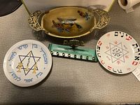 Top view of two ceramic Judaica plates, an oval brass tray, and a blue-green metal menorah holder