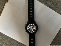 Black DT78 smart watch laid flat on white paper, showing the round face with analog clock display and two buttons on the right side of case.