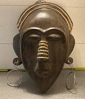 Front view of the first dark wooden elongated African mask with rope and metal hoop earrings, showing detailed carving and wrapped rope decoration