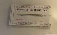 Front view of sealed plastic box labeled 'Stainless Steel Spring Bar' showing sizes available (9-24mm) and specifications including 1.50mm double flange diameter.