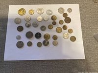 Top-down view of approximately 41 assorted world coins on a white sheet, showing different sizes and materials including silver, gold, and copper tones.