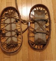 Top-down view of vintage bentwood bear claw snowshoes laid flat on a hardwood floor, showing the rope lacing and fabric bindings.