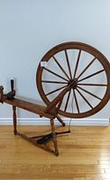 Full side view of the antique wooden spinning wheel showing the large wheel, seat, and legs on hardwood floor.