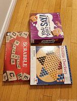 Photo showing three board games: Scrabble, You Don't Say, and Chinese Checkers, all boxed and stacked on a wooden floor.