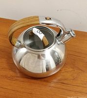 Stainless steel whistling kettle teapot with wood-look handle and lid knob on wooden surface.