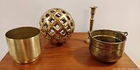 All four brass and brass-colored decorative items arranged on wooden surface: brass pot with handle, lattice ball decor, brass candlestick, and planter pot