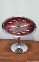 Front and side view of the oval-shaped Caravelle vintage alarm clock on tulip pedestal base, showing metallic red sunburst dial and chrome bezel.
