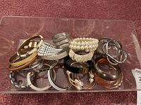 Clear plastic bin containing a variety of metal, beaded, and plastic bangle bracelets arranged inside.