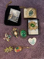 Nine assorted brooches arranged on a purple fabric background including floral, enamel, and stone styles with some in boxes and one in a black velvet pouch.