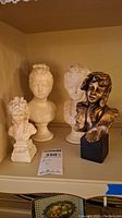 Four female bust sculptures on a shelf, three white busts and one bronze-tone bust, showing details and condition.