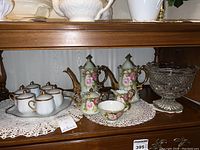 Full view of porcelain coffee pots with floral patterns, a teacup, and pressed glass bowl on a lace doily under a wooden shelf.
