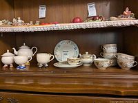 Photo showing several tea cups, saucers, plates and Carlton Ware teapot, creamer, sugar bowl on a wooden shelf with a lace shelf liner.