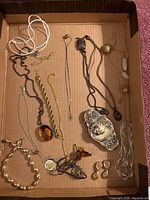 Full display of assorted costume jewelry laid inside a cardboard box, showing necklaces, earrings, and belt buckle.