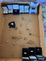 All costume rings arranged in a cardboard box, some in ring boxes and others loose, showing a variety of styles including gold-tone and silver-tone rings, some with simulated stones and adjustable bands.