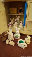 Full view of the lot laid out inside a cabinet and on carpet, showing multiple white ceramic rabbit figurines, one dressed rabbit figurine with fabric, and a yellow box behind them