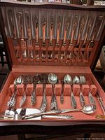 Full view of both flatware sets inside lined storage box with organized slots. Visible knives, forks, spoons, and serving pieces with floral patterned handles.