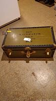 Top view of rectangular olive green military style trunk with black reinforced edges and stenciled text on top.