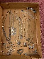 Multiple assorted jewelry pieces including necklaces, bracelets, earrings and pins shown arranged in a cardboard box.
