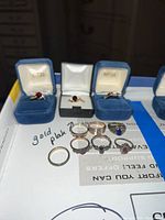 All 10 rings arranged on a surface with some in Avon blue ring boxes showing various styles and stone colors including red, purple, and blue.