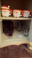 Ceramic Santa Claus mugs on top shelf with purple stem glasses and clear floral glasses below in cabinet.