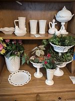 Shelf displaying various milk glass pieces including bowls, glasses, covered candy dish, and ceramic floral decorations.