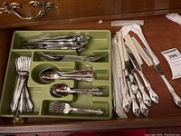 Wide view of flatware in green tray and loose bundled knives and forks on wood surface.