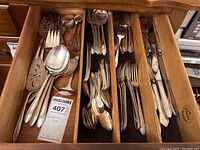 Top-down view into an open drawer showing a mix of silver-tone flatware and serving pieces with a wooden drawer organizer and a MaxSold lot tag for item 407 labeled 'Flatware Set'.