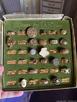 Top view of the original green box containing 36 assorted gold-tone rings in individual slots, showing a variety of designs including smooth, textured, and stone-set styles.