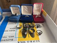 Ten sterling silver rings shown together, some in jewelry boxes labeled Avon and others in blue and red velvet boxes, displayed on a printed paper surface.