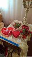 Full lot arranged on lace-covered table including a red heart-shaped tray with rope handles, white ceramic vase with hearts and artificial flowers, red heart-shaped ceramic pitcher, and other heart-shaped items.