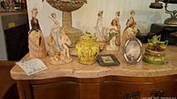 Wide view of 13 porcelain pieces including Victorian-style figures, trinket boxes, and framed photos on a marble top surface.