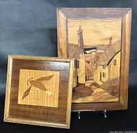 Two framed English marquetry wood panels shown together on a black background. The smaller panel features a bird motif, the larger a village street scene.