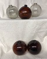 Five vintage glass fishing floats arranged on a white cloth, showing three floats in clear and amber colors (three floats top row, two floats bottom row)