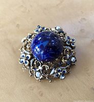 Front view of the brooch showing the deep blue faux lapis lazuli stone, blue enamel flowers, small faux pearls, and intricate brass filigree work.