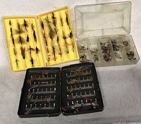 Photo showing two yellow plastic trays and one black metal box containing assorted fly fishing lures on a white surface.