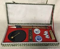Photo of vintage Chinese calligraphy set items in red felt-lined box including brushes, ink stick, ink stone with spoon, chop, and brush rests.
