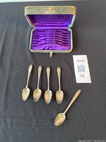 Photo showing 5 silver spoons with decorative handles and original storage case open above them with purple lining.