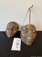 Shows both bronze masks side by side, one with an attached metal hanger, displayed on black fabric against white wall to illustrate size and condition.