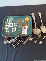 Overview of lot items on green tray with spoons and combs to right. Shows coins in protective holders, loose coins, vintage metal clamps, two large spoons, and small souvenir spoons.
